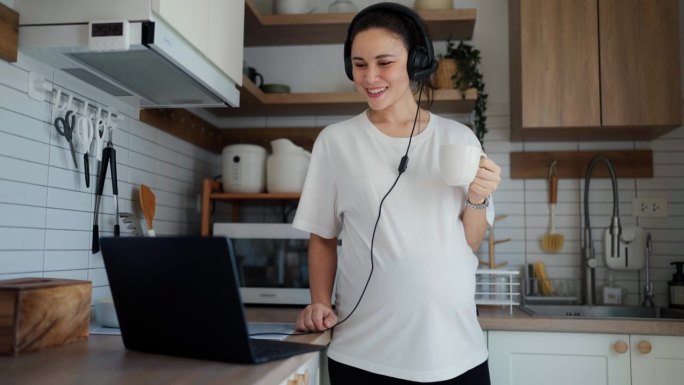快乐的亚洲孕妇站在家里的现代厨房里，喝着热牛奶，用笔记本电脑做网上工作