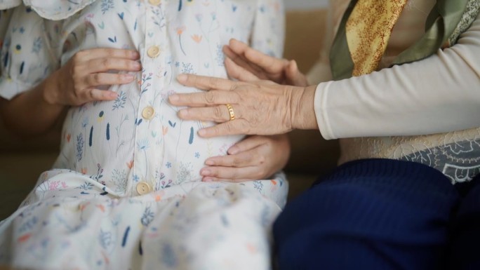 一位老妇人坐在家里的沙发上，用手抚摸着怀孕女儿的肚子。