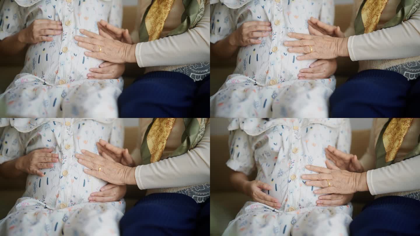 一位老妇人坐在家里的沙发上，用手抚摸着怀孕女儿的肚子。