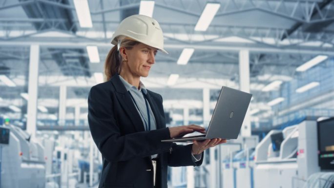 一个戴着安全帽的白人女工程师在电子制造工厂站着使用笔记本电脑的快乐画像。技术人员对工业机械任务和工作