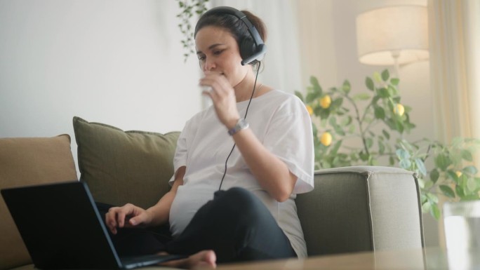 美丽快乐的孕妇坐在柔软的沙发上。在现代公寓里，用耳机和电脑工作