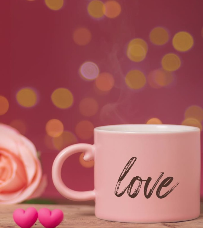 粉红色咖啡杯里用烟熏的热饮，旁边写着“爱”字，木桌上并排摆放着玫瑰和两颗小心，背景上散焦的散景光。浪