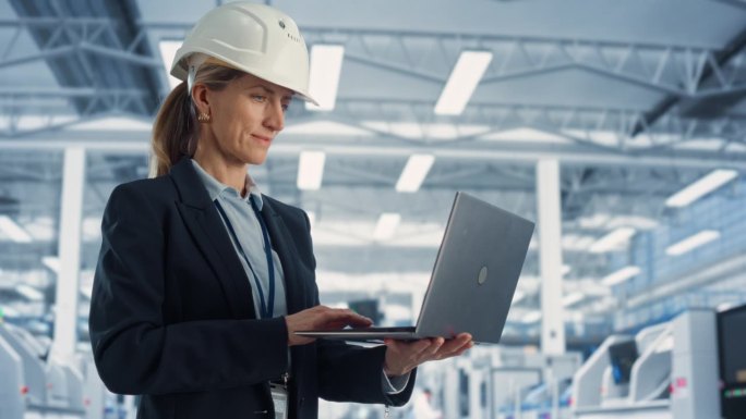 戴安全帽的白人女工程师在电子制造工厂站立和使用笔记本电脑的画像。技术人员思考日常任务并处理开发数据。