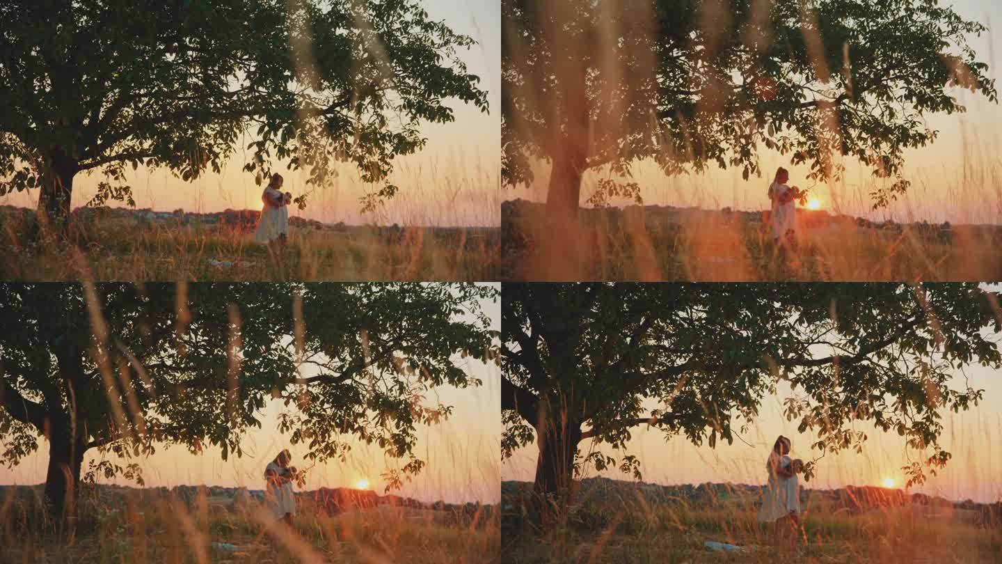 日落时分，满怀爱意的母亲抱着孩子在田野上的大树下背对天空的慢镜头镜头