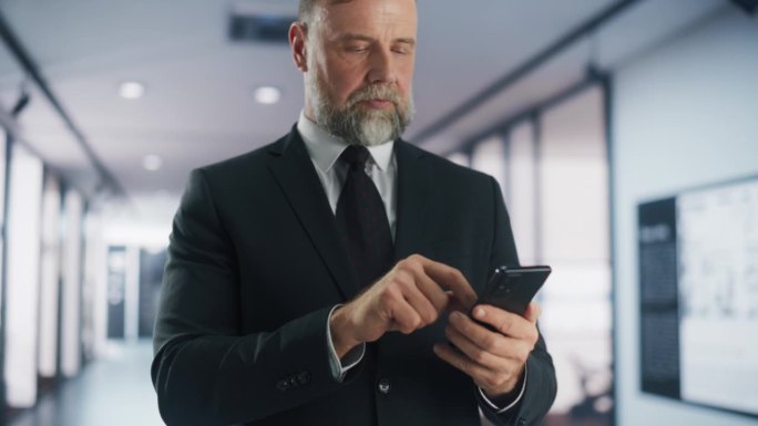 一个体贴的成年男子的肖像，穿着时髦的西装，拿着智能手机，微笑着站在公司办公室的走廊里。成功的商人用电