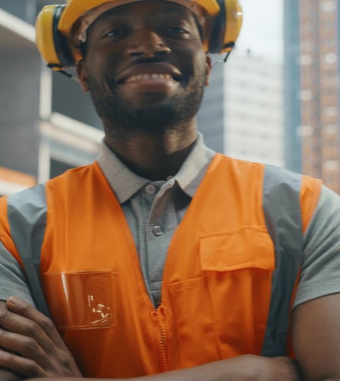 垂直屏幕:一个非洲建筑商的肖像，戴着安全帽和安全背心，在商业建筑工地摆姿势拍照和微笑。黑人交叉双臂
