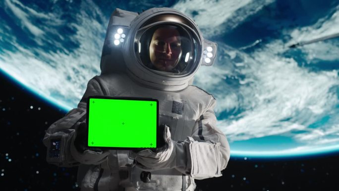 一名年轻的男性宇航员在卫星或空间站上进行太空行走时穿着宇航服摆姿势的肖像。宇航员手持平板电脑与绿色屏