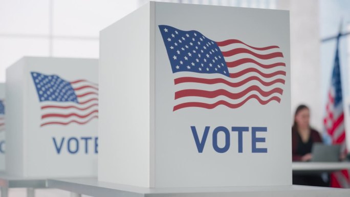 美国的选举日。投票站的桌子后面坐着一名匿名官员，投票站上有美国国旗的标志。为美国总统选举做准备