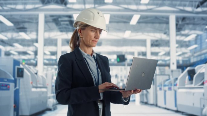 一位戴着白色安全帽的白人女性工业工程师站在电子制造工厂的笔记本电脑前，满脸笑容。成功的员工看着镜头。
