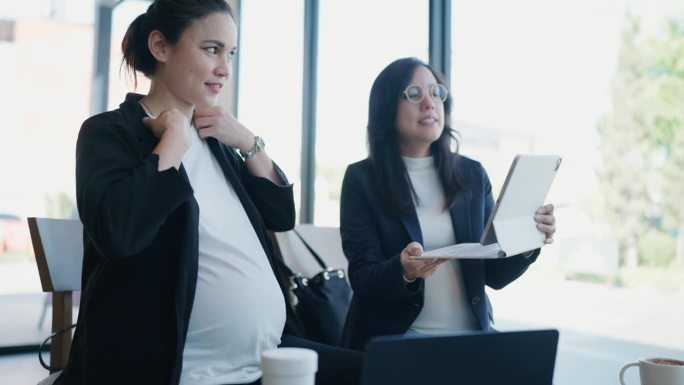 两个怀孕的亚洲妇女在咖啡馆用数码平板电脑讨论她的工作。