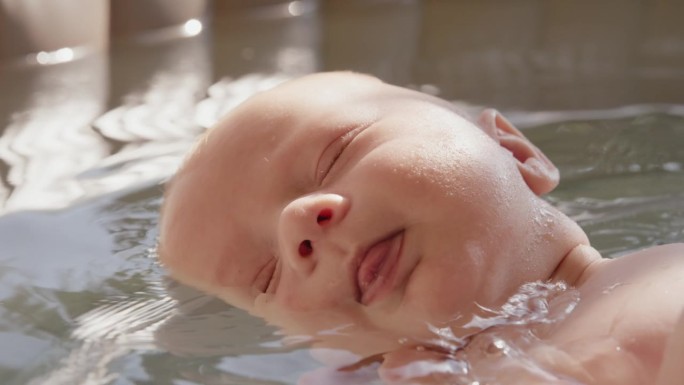 可爱的小男孩在浴缸里浮在水面上，妈妈的手在他的头下面
