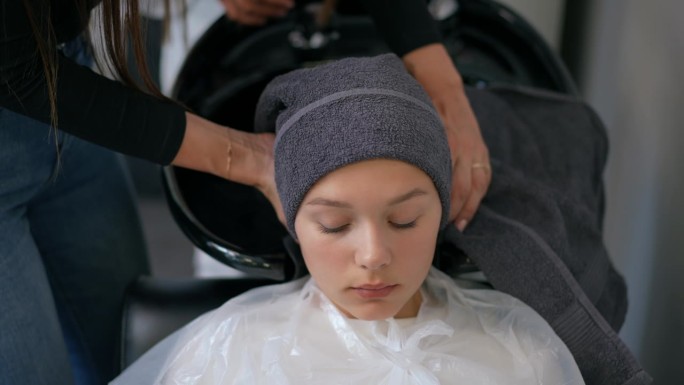 一位女理发师用毛巾擦干一个女孩的头发，把它缠在她的头上。女孩躺在理发师的椅子和水槽。