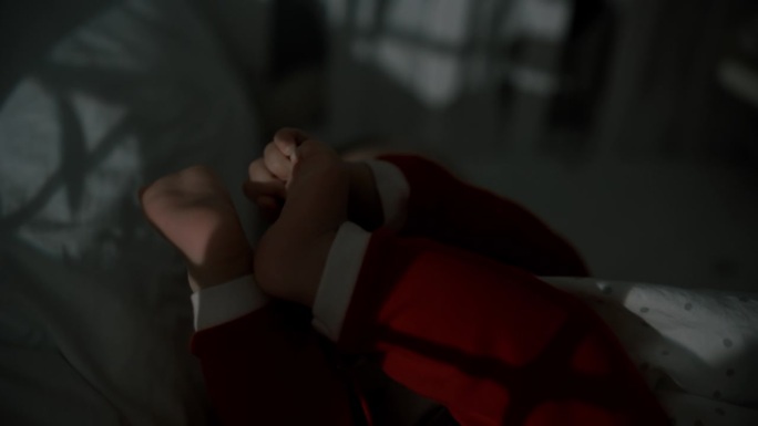 手持拍摄的顽皮的男婴在圣诞老人的服装躺在婴儿床在暗房白天