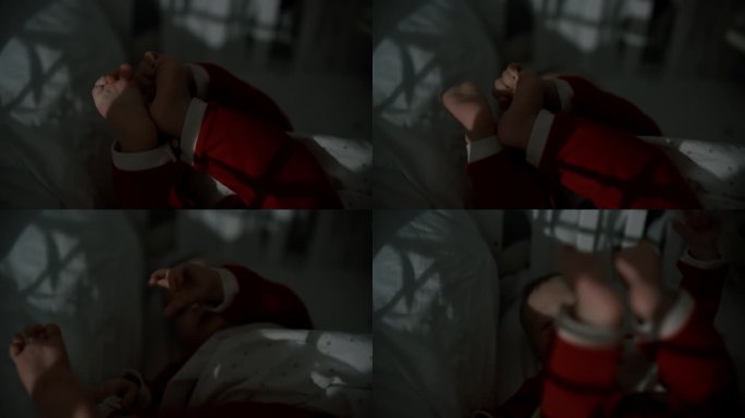 手持拍摄的顽皮的男婴在圣诞老人的服装躺在婴儿床在暗房白天