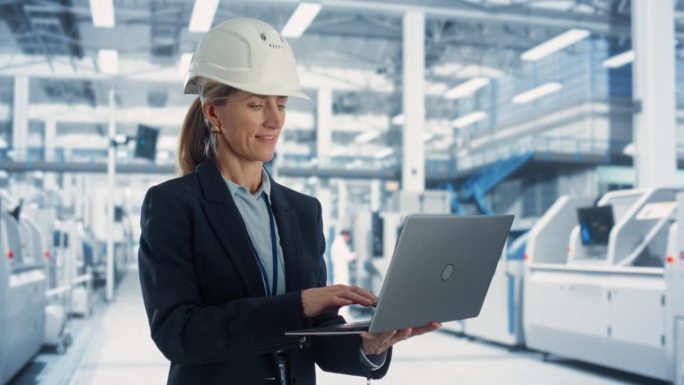 在一家重型机械制造工厂，戴着安全帽在笔记本电脑上工作的白人电子工厂女经理。专业监控生产，计划工作任务