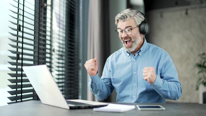 兴奋成熟的白发大胡子商人戴着无线耳机在公司办公室用笔记本电脑观看体育比赛
