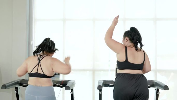两个胖乎乎的亚洲女人穿着运动服在跑步机上跑步。两个胖乎乎的女人都有强烈的减肥意愿、健身伙伴、生活方式