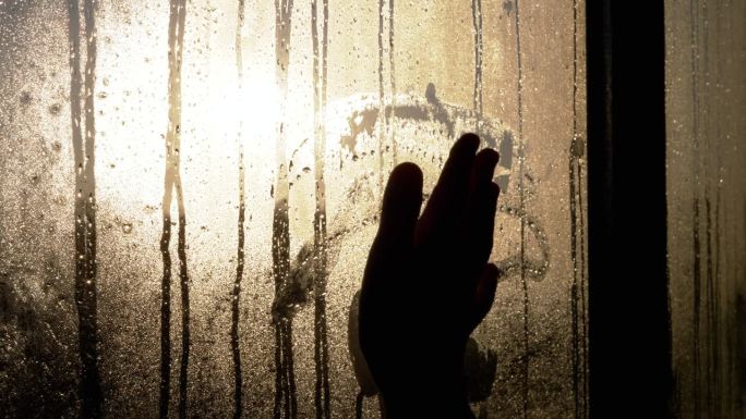 一个女人的手的剪影触摸雾蒙蒙的潮湿的窗户在闪烁的阳光眩光