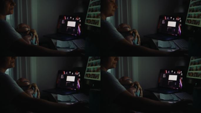 在黑暗的家庭办公室，母亲抱着熟睡的男婴使用台式电脑的SLO手持镜头。在家和宝宝一起工作。