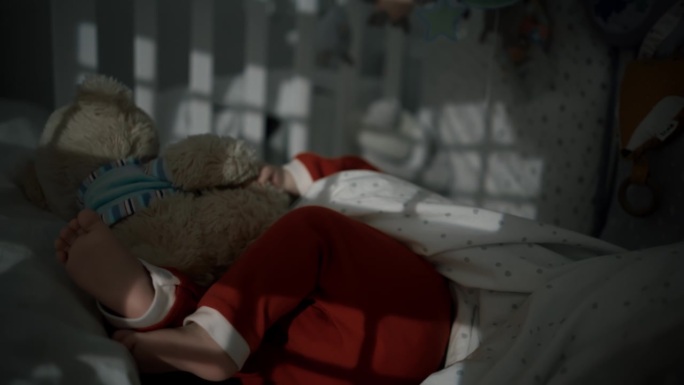在家暗房里，穿着圣诞老人服装的小男孩躺在婴儿床上和泰迪熊玩耍的手持照片