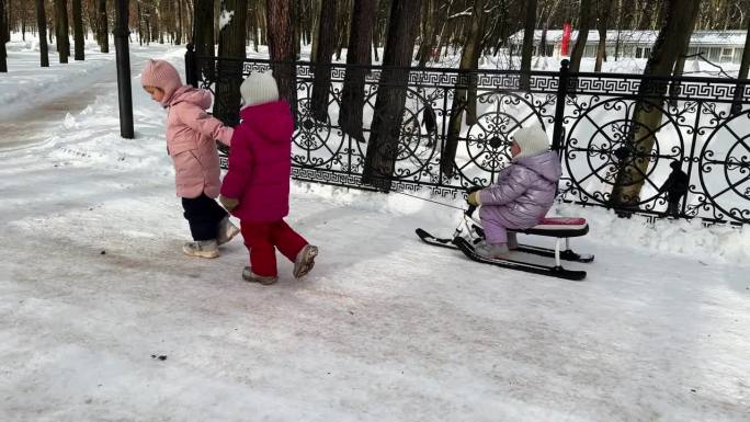 城市公园里，两个小妹妹用绳子拉着坐在雪橇上的第三个妹妹