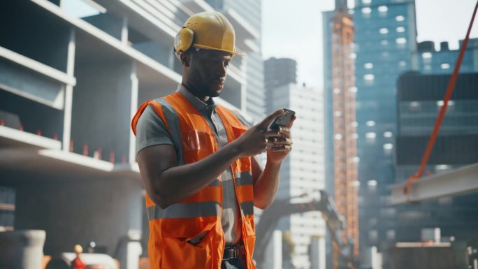英俊的非洲建造者、工人、工匠的肖像，戴着安全帽和背心，站在商业建筑工地，用智能手机浏览社交媒体