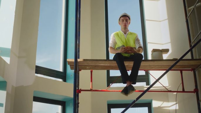 一个工人在建筑工地吃三明治，高高地坐在脚手架上
