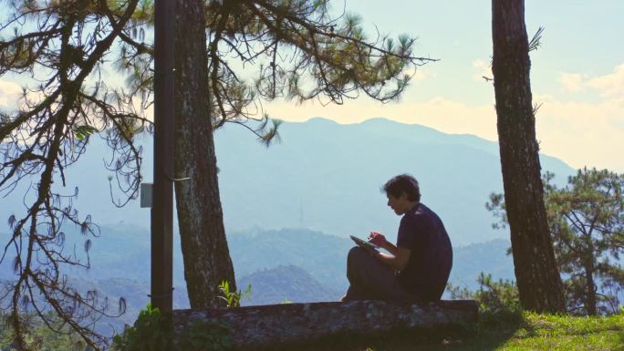 数字游牧民和旅行的概念与单独的自由职业者男人坐在木头和使用平板电脑工作从户外层山的背景