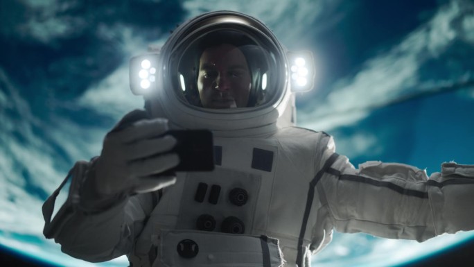 一个年轻的宇航员在零重力下漂浮在太空中的肖像。宇航员在轨道飞船外用智能手机观看视频。男子上网，连接卫