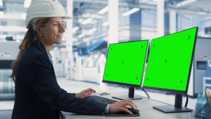 在工厂办公室里，戴安全帽的白人女技术人员在带有绿屏Chromakey模拟显示器的台式电脑上工作。自主