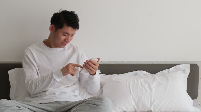 年轻帅气的中国男子在卧室的床上使用手机