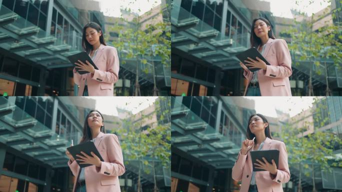 穿着漂亮的年轻亚洲女商人手持数码平板电脑，使用软件应用程序，站在高楼林立的街道上，女经理使用数码平板