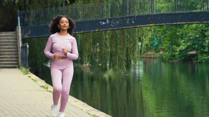 身穿粉色健身服的年轻女子沿着运河跑步穿过市区——慢镜头