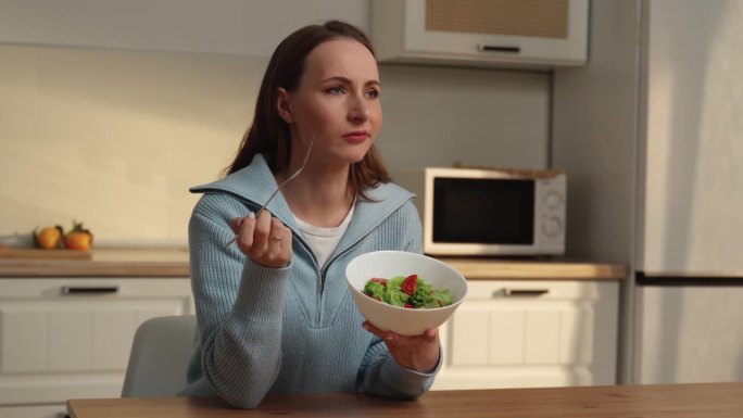 食欲不振。一个不开心的女人坐在厨房里，手里拿着叉子，端着一份蔬菜沙拉。失去味觉和嗅觉