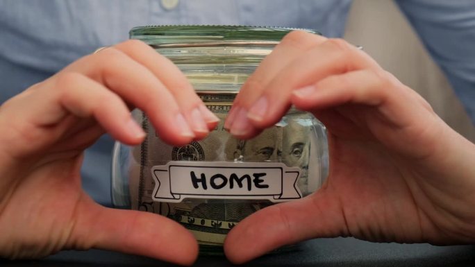 女性的双手在装满美国货币、美元、现金、纸币的玻璃罐前展示着爱心的标志，上面写着“家”。准备省钱。适度