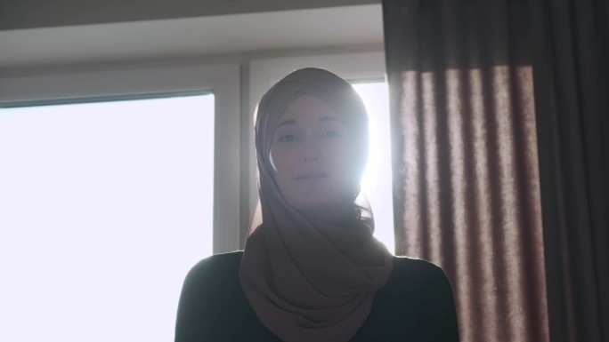 戴着头巾的穆斯林妇女站在房间的窗户附近。强光照进摄像机，使视频模糊不清。肖像。一个自信的女人的概念。