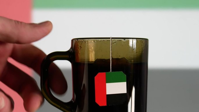 一个男人的手在玻璃桌子上放着一杯茶，茶包上有阿联酋国旗的标签，与同样模糊的国旗相对