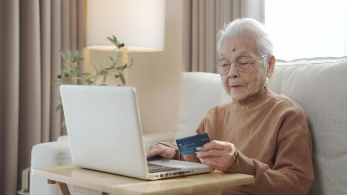 戴着眼镜、满头白发的老妇人坐在客厅里，手里拿着银行卡，用着智能手机