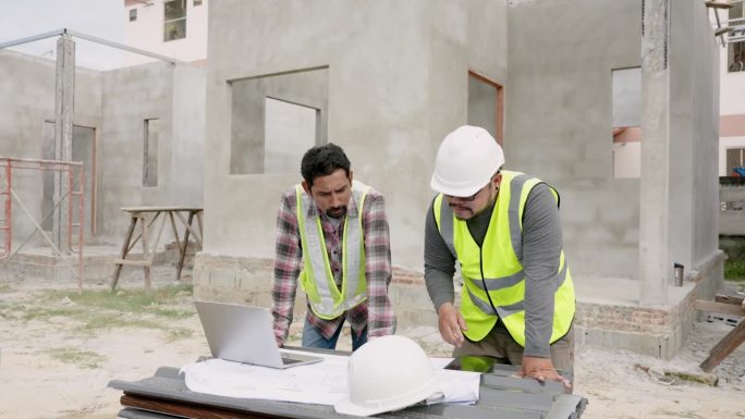现场工人讨论建筑方案。戴着白色头盔和黄色背心的亚洲工程师们用笔记本电脑和文件组成团队建造房屋。未完工