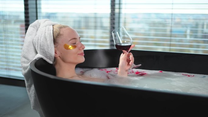 水疗中心放松。女人躺在浴室里，手里拿着一杯红酒。女孩在家里的浴室里放松。漂亮的女人在洗热水澡，喝红酒