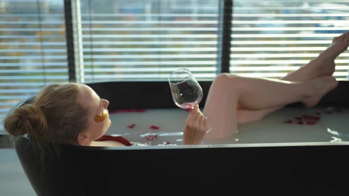 水疗中心放松。女人躺在浴室里，手里拿着一杯红酒。女孩在家里的浴室里放松。漂亮的女人在洗热水澡，喝红酒