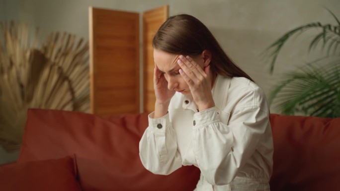 一个头痛得厉害的疲惫女人。患有急性偏头痛或颞动脉炎的少女正坐在家里的沙发上
