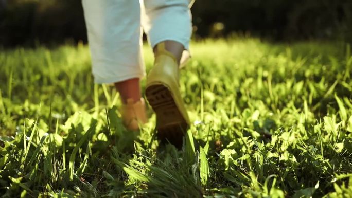 园丁脚踩黄色胶靴，走在绿草地和碎石地上。秋天的季节。特写镜头。从上面看。身体的部分。农夫在农场里走来