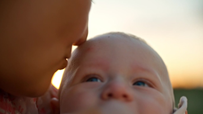 SLO - MO手持拍摄的剪短的母亲在日落时亲吻欢快的男婴额头的照片