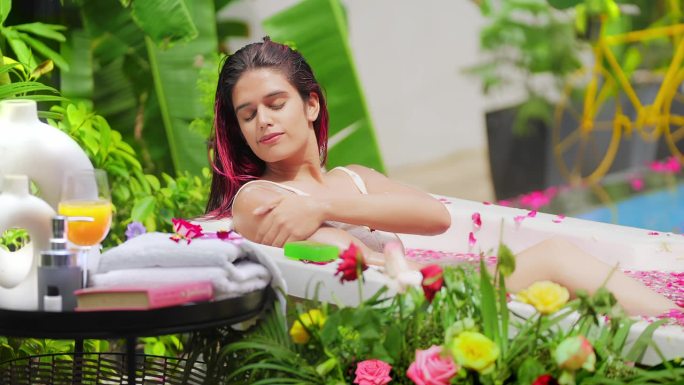 微笑的印度女人躺在浴缸里放松地用肥皂洗手。快乐的女性在酒店度假胜地沐浴清新的玫瑰花瓣水