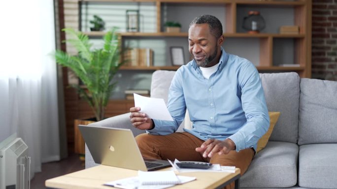 成熟的非裔美国男性坐在家里的沙发上用笔记本电脑支付水电费。