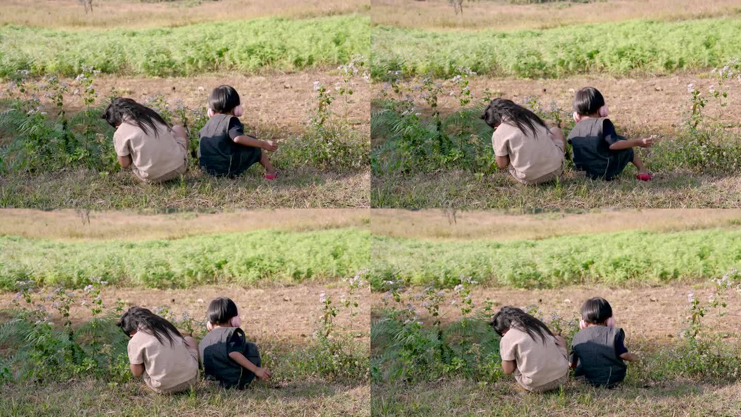 两个亚洲孩子背靠背坐在田野里，一个戴着耳机，另一个看着野花，和平共处。兄弟姐妹在户外分享安静的时刻，
