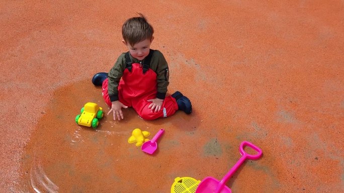孩子穿着防水夹克和防水靴子，在水坑里愉快地玩着他的玩具车。两岁男童(2岁)