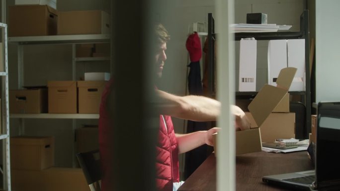 一名身穿红色背心的男子，是一名仓库员工，正在将包裹装入投递箱并关闭，他坐在纸板箱的背景上。物流、配送
