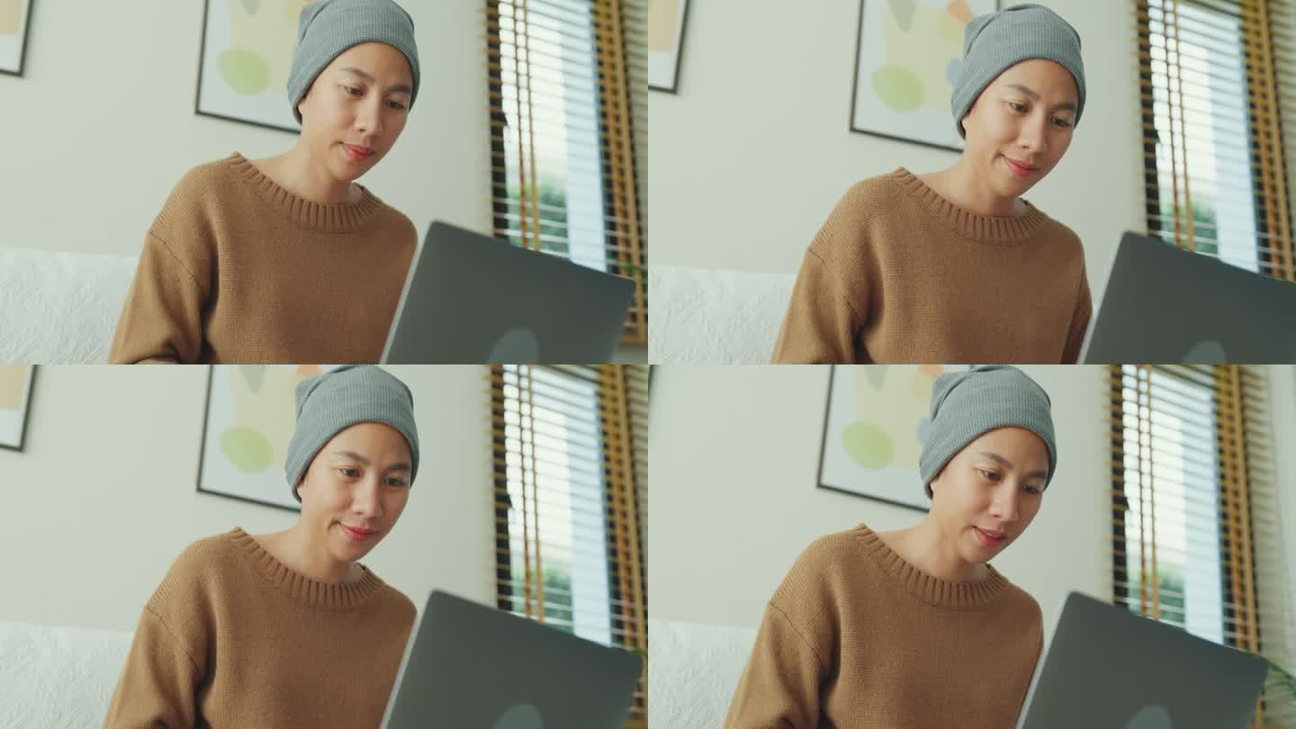 一个年轻的亚洲癌症患者，戴着头巾，舒服地坐在沙发上用笔记本电脑工作，房间里光线充足，植物装饰。与癌症
